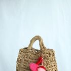 Geflochtene Tasche aus hellem Material mit buntem Anhänger aus roten und rosa Bommeln auf hellem Hintergrund.