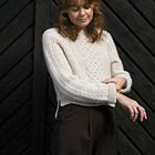 Braun-gemusterte Holzwand im Hintergrund; Person mit gelocktem Haar, trägt cremefarbenen Strickpulli und dunkle Hose.