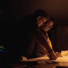 Zwei Personen arbeiten bei schwachem Licht an einem Tisch und studieren aufmerksam Dokumente unter einer Lampe.