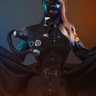 Futuristische Figur in schwarzem Latexanzug mit Schläuchen und Maske, neonblaues und orangenes Licht.