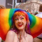 Eine Person mit einem breiten, regenbogenfarbenen Hut lächelt fröhlich. Ihr Make-up ist bunt, passend zum Hut.