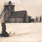 Eine Gruppe von Skifahrern steht bei einer kleinen Stein- und Holzhütte im Schnee, teilweise mit Ski ausgestattet.