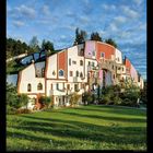 Zahlen von 1 bis 28 unten, mit „II 2027“. Ein Gebäude mit unregelmäßiger, bunter Fassade und viel Grün im Vordergrund.