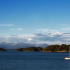 Ein weißes Segelboot auf einem blauen Meer vor bewaldeten Inseln und Bergen im Hintergrund, unter klarem Himmel.
