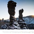 Kalenderblatt Februar. Schneebedeckte Felsformationen bei Sonnenuntergang, umgeben von Wäldern.