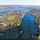 "Insel im Fluss" in großer Schrift. Luftaufnahme einer Stadt mit Fluss, Grünflächen und Booten.