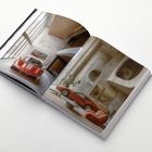 Ein aufgeschlagenes Buch zeigt luxuriöse Innenräume mit roten Sportwagen vor moderner Architektur und Möbeln.