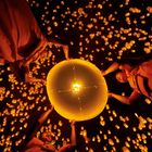 Menschen lassen eine große, leuchtende Papierlaterne steigen, umgeben von vielen kleinen schwebenden Lichtern in der Dunkelheit.