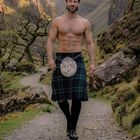 FEBRUAR. Ein muskulöser Mann in einem Kilt wandert auf einem Pfad in einer felsigen, grünen Landschaft.