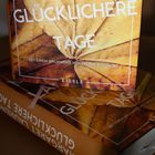 Ein Buch mit der Aufschrift "Laurence Roman Glücklichere Tage". Hintergrund: ein gelbes Ahornblatt.