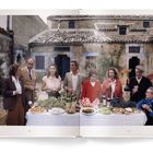Mehrere Menschen stehen an reich gedecktem Tisch mit Weinflaschen, Speisen und Trauben; im Hintergrund alte Gebäude.