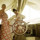 Zwei Frauen auf einem Segelboot. Eine steht in einem bunten Kleid, die andere lehnt am Steuerrad in einem weißen Kleid.