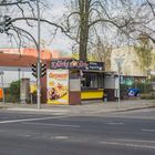 „Det is Knorke“, „Currywurst“, „Alte Berliner Currywurst-Spezialitäten“. Imbissbude an einer Straßenecke.