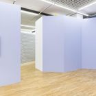 Links eine weiße Reliefskulptur auf lila Wand; rechts ein Podest mit transparentem Würfel; Holzboden.