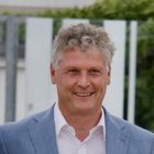 Es gibt keinen Text zu beachten. Ein Mann mit Locken lächelt und trägt ein hellblaues Sakko und ein weißes Hemd.