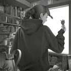 Eine Person mit Dutt betrachtet eine Erdbeere am Fenster, umgeben von Bücherregalen und Fruchtkörben in einem gemütlichen Raum.
