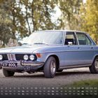 Text: "1 2 3 4 ... 28 Mo Di Mi Do Fr Sa So ... BMW E3 2." Ein klassisches blaues Auto steht auf einem Kiesweg.