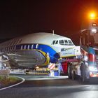 Zwei Lastwagen transportieren nachts einen großen Flugzeugrumpf auf der Autobahn. Lichter leuchten hell.