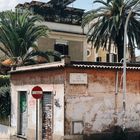 Straßenschild mit "VIA CINO DA PISTOIA", alte Hausfassade, Palmen im Hintergrund, Stoppschild vorne.
