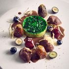 Grünes Dessert, umgeben von Datteln, Beeren und Schokoladensauce, auf einem weißen Teller kunstvoll angerichtet.