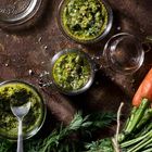 Ein Glas mit Pesto, umgeben von frischen Karotten mit Grün, auf einer rustikalen, braunen Oberfläche.