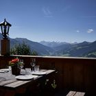 Ein Holzterrassentisch ist eingedeckt mit Gläsern und Tellern, umgeben von Alpenlandschaft und klarem Himmel.