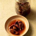 Glas mit dunklem Inhalt, Löffel darin. Teller mit aufgeschnittener Feige und Flüssigkeit, beige Untergrund.