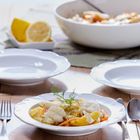 Ein Tisch mit weißen Tellern, Gabeln und Löffeln. Im Vordergrund ist ein Teller mit Fisch, Gemüse und Dill. Im Hintergrund liegt eine halbe Zitrone und eine Schüssel mit einem Löffel darin.