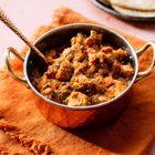 Ein Kupfergefäß mit Curry, serviert auf einem orangefarbenen Stoff. Rechts im Hintergrund sind Fladenbrote zu sehen.