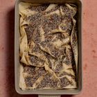 Kein Text vorhanden. Eine rechteckige Backform mit zerknittertem Teig, bestreut mit Mohn, auf rosafarbenem Untergrund.