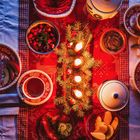 Ein festlich gedeckter Tisch mit roten und weißen Mustern, Kerzen und Tannenzweigen; umgeben von Speisen und Getränken.