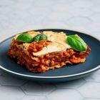Ein Teller mit einer Lasagne, dekoriert mit Basilikumblättern, auf einem blauen Teller auf einem hellen Tisch.