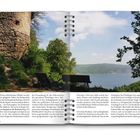 Ein großer, runder Turm aus Stein neben einem Rad und einer Bank am See mit Wald im Hintergrund. Blauer Himmel, klare Sicht.