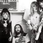 "Fleetwood Mac" steht auf einer Tafel im Hintergrund. Fünf Personen in Retro-Kleidung; eine Person hält eine Gitarre.