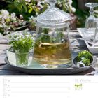 "Waldmeisterbowle auf einem dekorierten Tablett". Kalender mit Wochentagen im April 2027. Glasgefäß mit Kräutern, Frühlingsblumen.