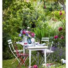 19. Mai: Ein weißer Tisch im Garten, gedeckt mit bunten Blumen, umgeben von grüner Vegetation und blühenden Sträuchern.