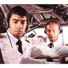 Zwei Piloten in Uniform mit Kopfhörern in einem Cockpit. Der vordere konzentriert, der hintere schaut direkt. Instrumente umgeben sie.