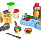 Text: "Color", "Okayers". 
Spielset mit Knete und Zubehör, darunter bunte Behälter und ein Plastikmesser.