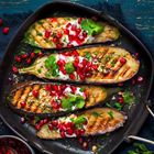 Gegrillte Auberginen auf einer Grillpfanne, garniert mit Joghurt, Granatapfelkernen und frischen Kräutern.