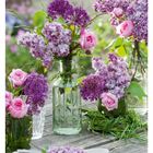 22 MAI/JUN. Ein Tisch mit Vasen voller Blumen: Flieder, Rosen und Zierlauch in lila und pinken Tönen.