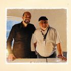 Zwei lächelnde Männer stehen nebeneinander an einem Tisch in einem hellen Raum. Der Mann rechts trägt eine Mütze.