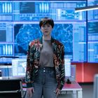 Mehrere Computerbildschirme mit Daten. Eine Person steht im Vordergrund, trägt eine bunte Jacke und Jeans im Büro.