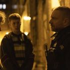 Text auf Uniform: "POLICE". Drei Personen in dunkler Straße, warmes gelbes Licht wirkt nächtlich und konzentriert.
