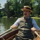 Ein Mann in altmodischer Kleidung rudert auf einem Fluss. Im Hintergrund ist eine Steinbrücke und bewaldete Ufer.