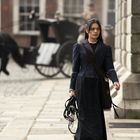 Eine Frau in schwarzer Kleidung geht einen gepflasterten Weg entlang, hinter ihr eine Kutsche und ein schwarzes Pferd.