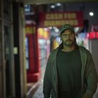 Im Hintergrund steht "CASH FOR DIAMONDS". Ein Mann mit Brille und Cap geht abends durch eine belebte Straße.