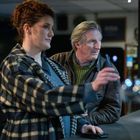 Eine Frau in kariertem Mantel und ein Mann stehen an einer Theke in einer Bar.