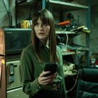 Eine Frau hält ein Smartphone in der Hand, steht in einem unordentlichen Raum mit Regalen und Kegel-Pins im Hintergrund.