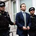 Zwei Polizisten in Uniform begleiten einen eleganten Mann in Anzug und Krawatte.