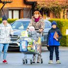 Ein Einkaufswagen mit Oki Einlagen und verschiedenen Lebensmitteln. Drei Personen stehen auf einer Straße vor einem Haus.